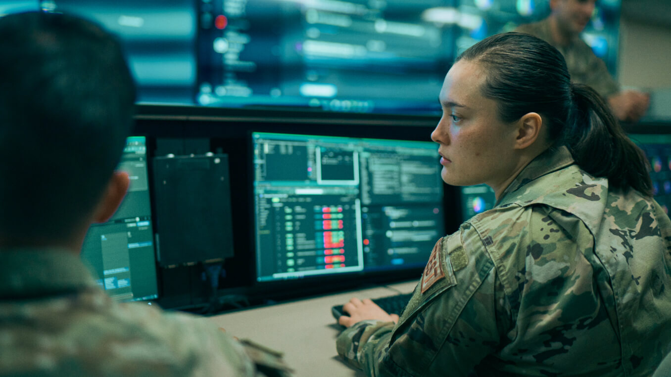 Two uniformed Cyberspace Warfare Operations service members review data on computer screens. 