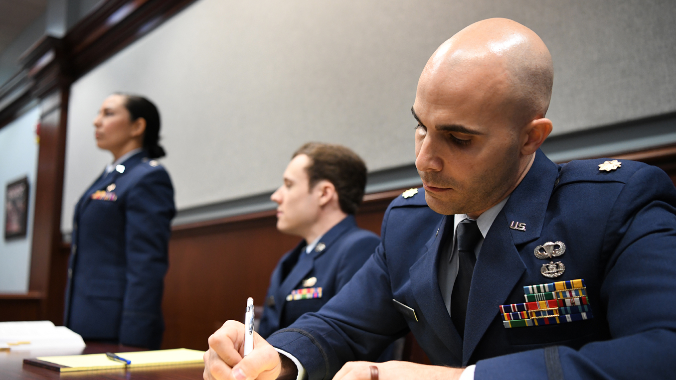 jag attorney writing on a notepad while two other JAG attorneys pay attention in court